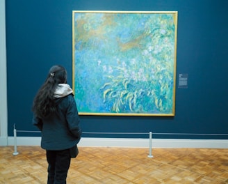 A person stands in front of a large artwork in a museum, observing an impressionist painting that features a blend of blues, greens, and hints of yellow. The painting is displayed on a dark blue wall with a simple, elegant frame. The floor is wooden, enhancing the serene environment.