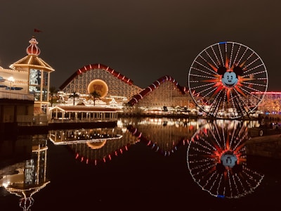 A nighttime amusement park scene with a large Ferris wheel featuring an iconic character's face, surrounded by bright lights. The structure is reflected in a body of water, creating a mirror image effect. The park features a roller coaster with bright red tracks and a lit-up pavilion on the left side.