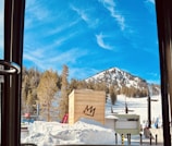 A snow-covered mountain under a bright blue sky with scattered white clouds. Pine trees line the mountain slope, and a wooden crate with a stylized crown symbol sits in the foreground. Skiers can be seen in the background on the ski slopes.