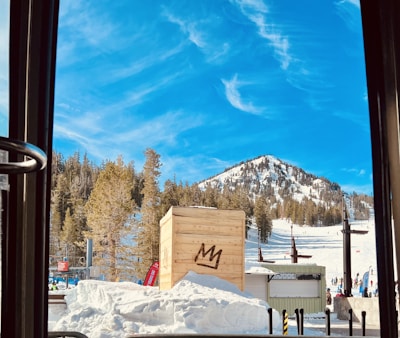 A snow-covered mountain under a bright blue sky with scattered white clouds. Pine trees line the mountain slope, and a wooden crate with a stylized crown symbol sits in the foreground. Skiers can be seen in the background on the ski slopes.