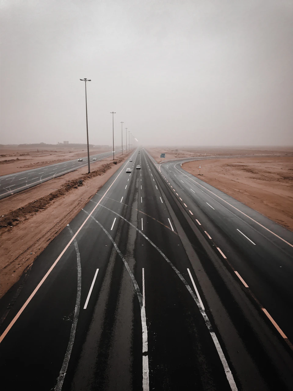 The image depicts a long, straight highway stretching into the distance with two lanes filled with vehicles moving in opposite directions. The sky above is hazy and overcast, casting a soft light on the scene below. In the background, there are several buildings visible at the edge of the highway, suggesting that this road may be located near an urban area or town.

The perspective of the image is from above, providing a bird's eye view of the highway and its surroundings. The overall color palette is dominated by shades of brown and gray, reflecting the hazy sky and the earthy tones of the desert landscape.