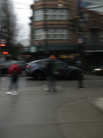 A street scene with a blurred motion effect. A dark-colored car is moving quickly past a city intersection, surrounded by blurred pedestrians. The background features a multi-story brick building with a distinct architectural design and a partially visible green sign.