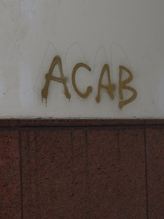 Graffiti with the letters ACAB is sprayed in brown on a light-colored wall. Below the graffiti, there is a dark-colored tiled surface.