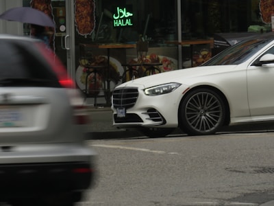 A white luxury car is parked on a city street near a restaurant with a neon sign that reads 'Halal'. Another car is partially visible, driving past, and a person holding an umbrella is walking on the sidewalk.