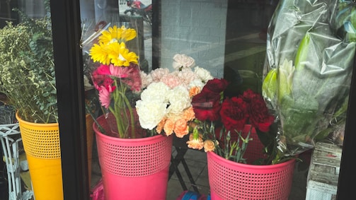 A collection of vibrant flowers displayed in plastic baskets next to a glass window. The assortment includes red, pink, white, and yellow blossoms, with some wrapped in plastic. The setting appears to be indoors, likely a flower shop, with a brick wall and reflections of the outside street visible.