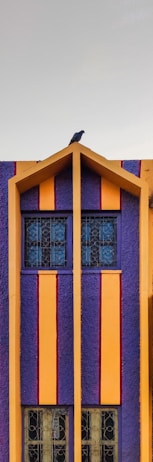 A vibrant building facade featuring bold vertical stripes in purple and orange with a small triangular roof section. A bird perches at the top, adding a touch of nature to the geometric design. The window frames and decorative patterns on the windows complement the modern, colorful aesthetic.