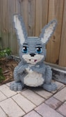 A gray and white bunny made out of LEGO bricks is placed on a tiled surface against a wooden fence. The bunny has expressive blue eyes and large ears, with a small pink tongue protruding from its mouth. The setting includes some greenery and mulch beside the tiles.