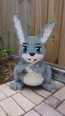 A gray and white bunny made out of LEGO bricks is placed on a tiled surface against a wooden fence. The bunny has expressive blue eyes and large ears, with a small pink tongue protruding from its mouth. The setting includes some greenery and mulch beside the tiles.