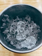 A metal cup filled with several clear ice cubes, set on a light wooden surface.