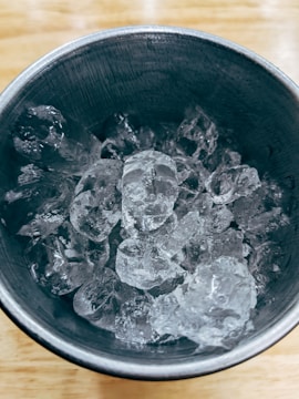 A metal cup filled with several clear ice cubes, set on a light wooden surface.
