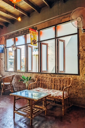 A cozy interior with rustic elements, featuring a rattan sofa and glass-topped coffee table. The walls are made of exposed brick, creating a warm and vintage atmosphere. Several wooden-framed windows allow soft light to enter, adorned with decorative banners. The seating area is complemented by a potted plant and wicker chairs.