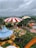 A vibrant amusement park scene with colorful tents, including a large red and white striped circus tent and a yellow and white striped tent. There are landscaped areas with pathways, greenery, and various themed structures. In the center, a small roundabout ride with animal figures surrounded by a fence is visible, and several people are walking around the park. The sky is overcast, creating a contrast with the bright colors below.