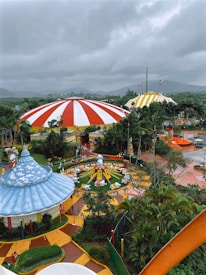 A vibrant amusement park scene with colorful tents, including a large red and white striped circus tent and a yellow and white striped tent. There are landscaped areas with pathways, greenery, and various themed structures. In the center, a small roundabout ride with animal figures surrounded by a fence is visible, and several people are walking around the park. The sky is overcast, creating a contrast with the bright colors below.
