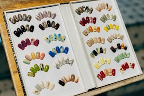 A display book featuring a variety of artificial nails with different designs and colors. The left side shows nails with a range of artistic patterns, including glitter, matte, and vibrant hues like pink, blue, and green. The right side presents more designs, including floral prints, geometric patterns, and glittering finishes. The nails are neatly organized in rows increasing the visual appeal of the collection.