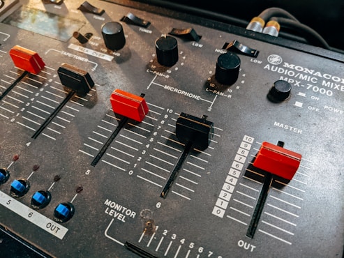 A close-up view of an audio mixer, featuring several sliders and knobs used for adjusting sound levels. The sliders are marked with numeric levels, including sections labeled 'Microphone' and 'Master'. The mixer is labeled 'Monacor Audio/Mic Mixer MPX-7000'.
