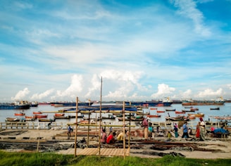 A coastal scene with small fishing boats anchored near a dock, where people are working with fishing nets. The background features large cargo ships on a vast expanse of calm water under a partly cloudy sky. The area appears lively with activity and maritime operations.