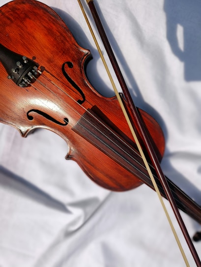 A wooden violin resting on a white fabric, accompanied by a bow. The body of the violin has a rich, warm brown color with visible wood grain. The bridge, strings, and scroll are clearly visible.