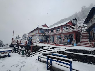 A snowy scene featuring a two-story building with colorful prayer flags draped across the front. The structure is painted red with white trim and has a staircase leading up to it. Snow covers the ground, steps, and rooftops, creating a serene and cold atmosphere.
