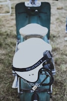 A vintage-style scooter with a brown leather double seat is parked on grassy ground. A white helmet with the inscription 'HIGHWAY 1' is placed on the seat, secured by a chain and padlock to the frame of the scooter. The scooter has a retro design with a teal body and chrome accents.