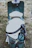 A vintage-style scooter with a brown leather double seat is parked on grassy ground. A white helmet with the inscription 'HIGHWAY 1' is placed on the seat, secured by a chain and padlock to the frame of the scooter. The scooter has a retro design with a teal body and chrome accents.