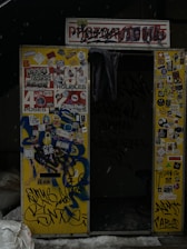 An old photo booth with a worn sign and partially obscured by graffiti. The exterior is covered in various stickers, drawings, and colorful graffiti including phrases and designs. The interior appears dark, with a curtain hanging inside. Snow is visible on the ground in front.