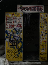 An old photo booth with a worn sign and partially obscured by graffiti. The exterior is covered in various stickers, drawings, and colorful graffiti including phrases and designs. The interior appears dark, with a curtain hanging inside. Snow is visible on the ground in front.
