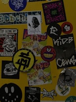 A collection of various stickers arranged on a yellow surface. The stickers include images of a woman, abstract designs, brand logos, and cartoon characters, along with text in different fonts.
