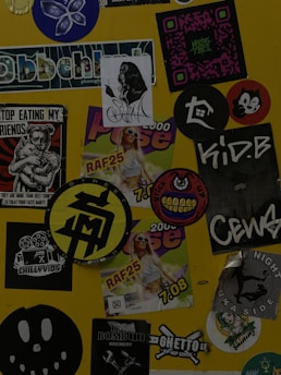 A collection of various stickers arranged on a yellow surface. The stickers include images of a woman, abstract designs, brand logos, and cartoon characters, along with text in different fonts.