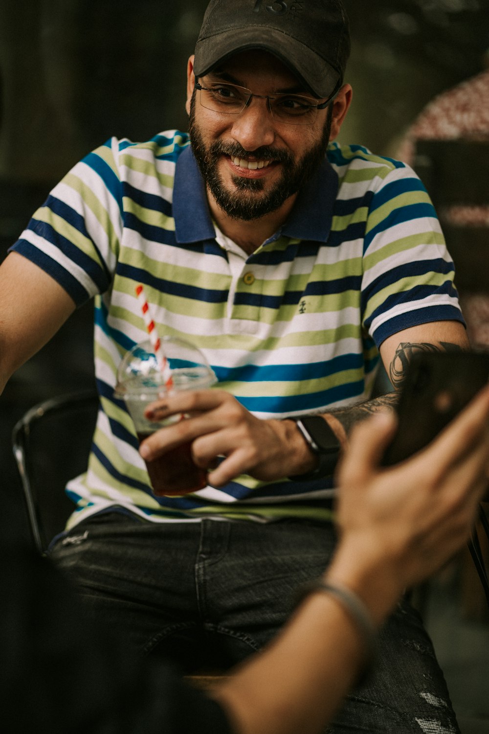 a man in a striped shirt and a black cap smiles as he holds a drink
