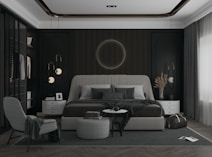 A modern bedroom with a large upholstered bed as the centerpiece, featuring dark paneled walls and minimalistic design elements. The room is decorated with pendant lights on either side of the bed, a circular wall decoration above the bed, and matching side tables. An armchair and a small round table with a book and coffee cup are placed near the bed, and there is a travel bag on the floor. The overall aesthetic is sleek and sophisticated with a hint of luxury.
