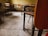 An empty classroom with wooden desks and chairs arranged in rows. The flooring is tiled, and the walls have a worn, yellowish hue. There is a calm and quiet atmosphere due to the absence of people.