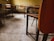 An empty classroom with wooden desks and chairs arranged in rows. The flooring is tiled, and the walls have a worn, yellowish hue. There is a calm and quiet atmosphere due to the absence of people.