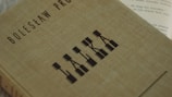 A hardcover book with a textured, beige cover features the name Bolesław Prus and the title 'Lalka' in a stylized, geometric font. The book is positioned diagonally in the frame, and part of an open book with visible text is partially seen in the background.