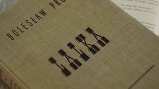 A hardcover book with a textured, beige cover features the name Bolesław Prus and the title 'Lalka' in a stylized, geometric font. The book is positioned diagonally in the frame, and part of an open book with visible text is partially seen in the background.