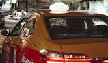 A yellow and red taxi with the word 'TAXI' illuminated on its roof drives through a city street at night. The vehicle's windows have visible raindrops, and the back seat is occupied by a passenger. City lights and blurred images of other vehicles are visible in the background, creating a busy nighttime atmosphere.