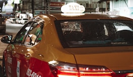 A yellow and red taxi with the word 'TAXI' illuminated on its roof drives through a city street at night. The vehicle's windows have visible raindrops, and the back seat is occupied by a passenger. City lights and blurred images of other vehicles are visible in the background, creating a busy nighttime atmosphere.