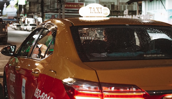 A yellow and red taxi with the word 'TAXI' illuminated on its roof drives through a city street at night. The vehicle's windows have visible raindrops, and the back seat is occupied by a passenger. City lights and blurred images of other vehicles are visible in the background, creating a busy nighttime atmosphere.