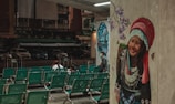 A mural depicting a smiling woman in traditional attire adorns a wall, with purple flowers and butterflies surrounding her. In the foreground, rows of green waiting area chairs are mostly empty, except for a few people sitting and interacting. The background reveals a dark, deserted street with parked cars and old buildings.