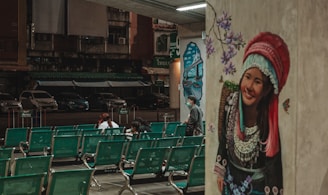 A mural depicting a smiling woman in traditional attire adorns a wall, with purple flowers and butterflies surrounding her. In the foreground, rows of green waiting area chairs are mostly empty, except for a few people sitting and interacting. The background reveals a dark, deserted street with parked cars and old buildings.