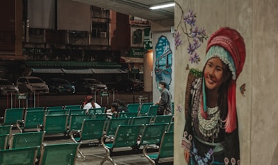 A mural depicting a smiling woman in traditional attire adorns a wall, with purple flowers and butterflies surrounding her. In the foreground, rows of green waiting area chairs are mostly empty, except for a few people sitting and interacting. The background reveals a dark, deserted street with parked cars and old buildings.