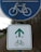 A blue circular road sign displays a white bicycle icon, indicating a bicycle lane or path. Below it, a rectangular sign features a green bicycle icon with an upward-pointing arrow, suggesting a direction for cyclists. The background includes a winding road, vegetation, and cloudy skies.