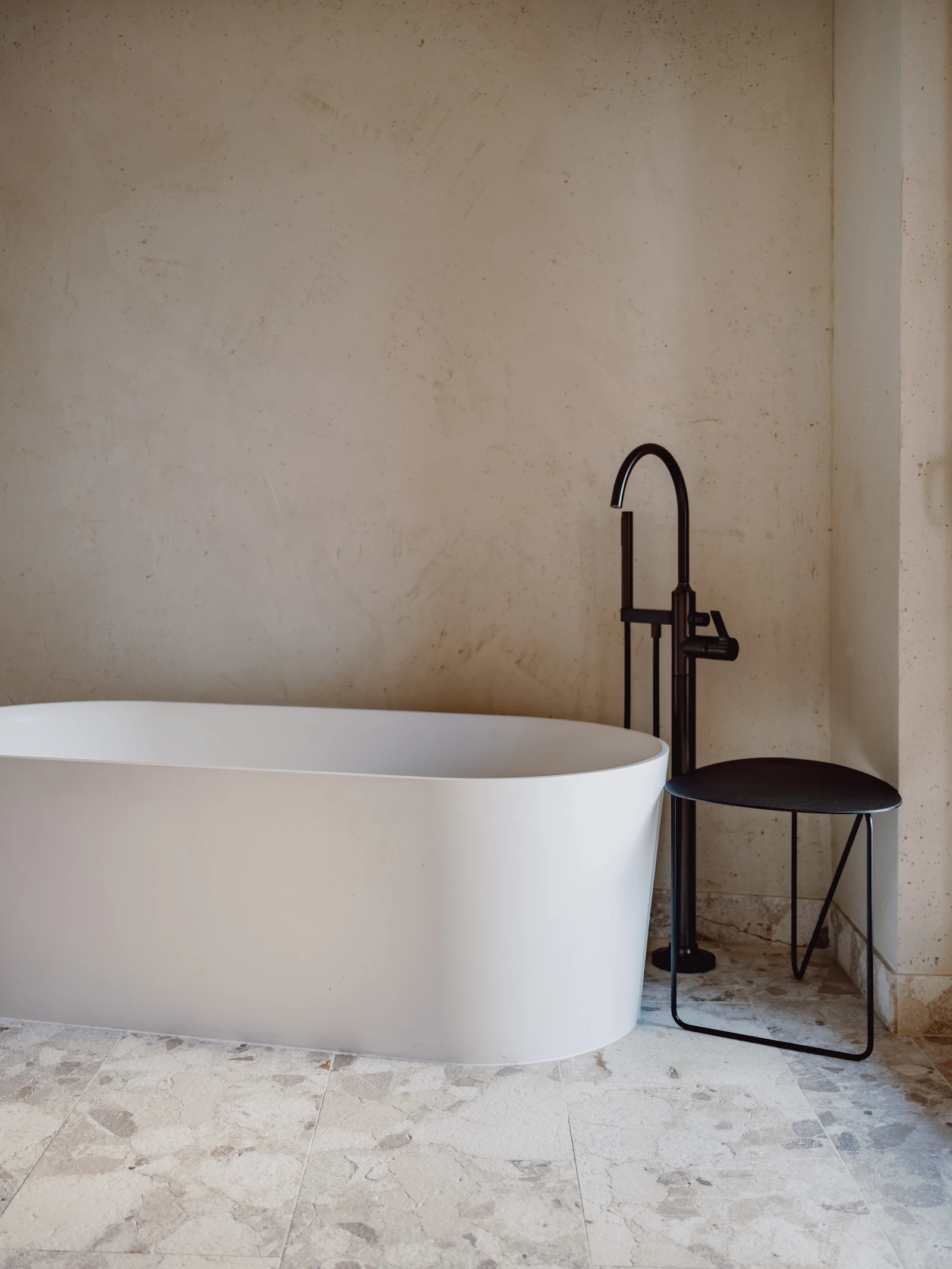 Minimalist bathroom with a freestanding white bathtub, dark sculptural floor-mounted tap, black chair, pale textured walls and patterned stone flooring.