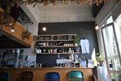 A cozy café interior with a menu board on a dark brick wall, shelves stocked with coffee-related items and a hanging white t-shirt. Plants are placed decoratively around the counter and windows, adding a touch of greenery. Four different colored chairs are aligned along the counter, and warm lighting creates an inviting atmosphere.