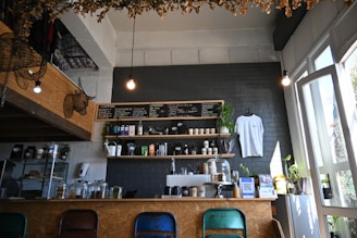 A cozy café interior with a menu board on a dark brick wall, shelves stocked with coffee-related items and a hanging white t-shirt. Plants are placed decoratively around the counter and windows, adding a touch of greenery. Four different colored chairs are aligned along the counter, and warm lighting creates an inviting atmosphere.