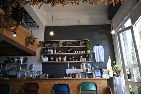 A cozy café interior with a menu board on a dark brick wall, shelves stocked with coffee-related items and a hanging white t-shirt. Plants are placed decoratively around the counter and windows, adding a touch of greenery. Four different colored chairs are aligned along the counter, and warm lighting creates an inviting atmosphere.