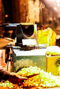 A street vendor's setup with a popcorn machine emitting steam. The scene is warmly lit, with fresh popcorn scattered around. A hand is visible, holding a tool or ladle to serve the popcorn. A yellow bag and other containers are in the background, suggesting a street market environment.