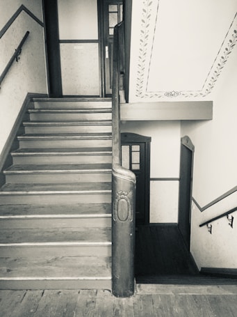 A vintage wooden staircase with decorative accents. The stairway features a handrail and ornate newel post, leading to a landing with access to multiple doors. The walls are adorned with a simple, elegant stencil pattern.