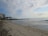 A sandy beach stretches along the shoreline with gentle waves lapping at the shore. To the left, there is a multi-story building with balconies, positioned near a cluster of palm trees. The sky is partly cloudy with patches of blue visible.