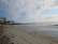 A sandy beach stretches along the shoreline with gentle waves lapping at the shore. To the left, there is a multi-story building with balconies, positioned near a cluster of palm trees. The sky is partly cloudy with patches of blue visible.