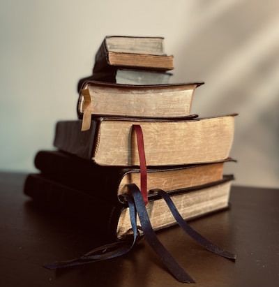 A stack of old hardcover books with visible signs of wear and textured covers. The books have attached fabric bookmarks in various colors, draped over the sides. The lighting casts soft shadows, giving a warm and nostalgic atmosphere.